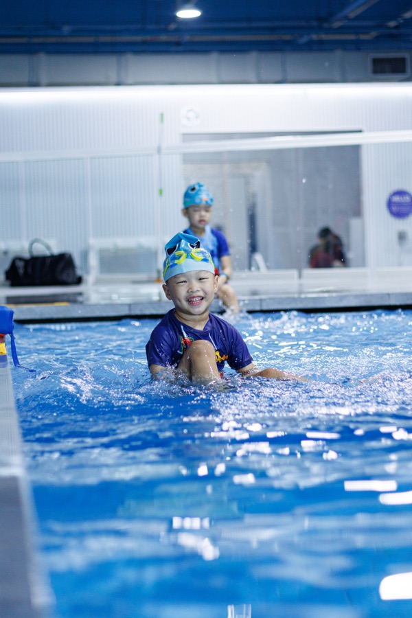 A toddler in a swim cap standing chest-deep in a lesson pool, looking at the coach