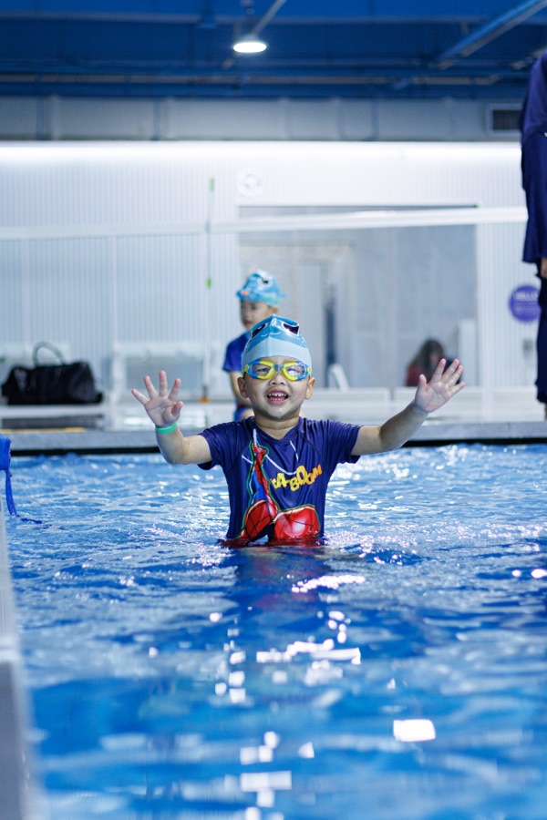 A toddler in a swim cap with both arms up in the water during a lesson